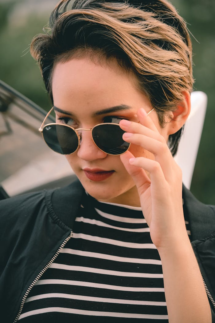 Fashionable woman with short brown hair adjusts her sunglasses outdoors. Captivating portrait.